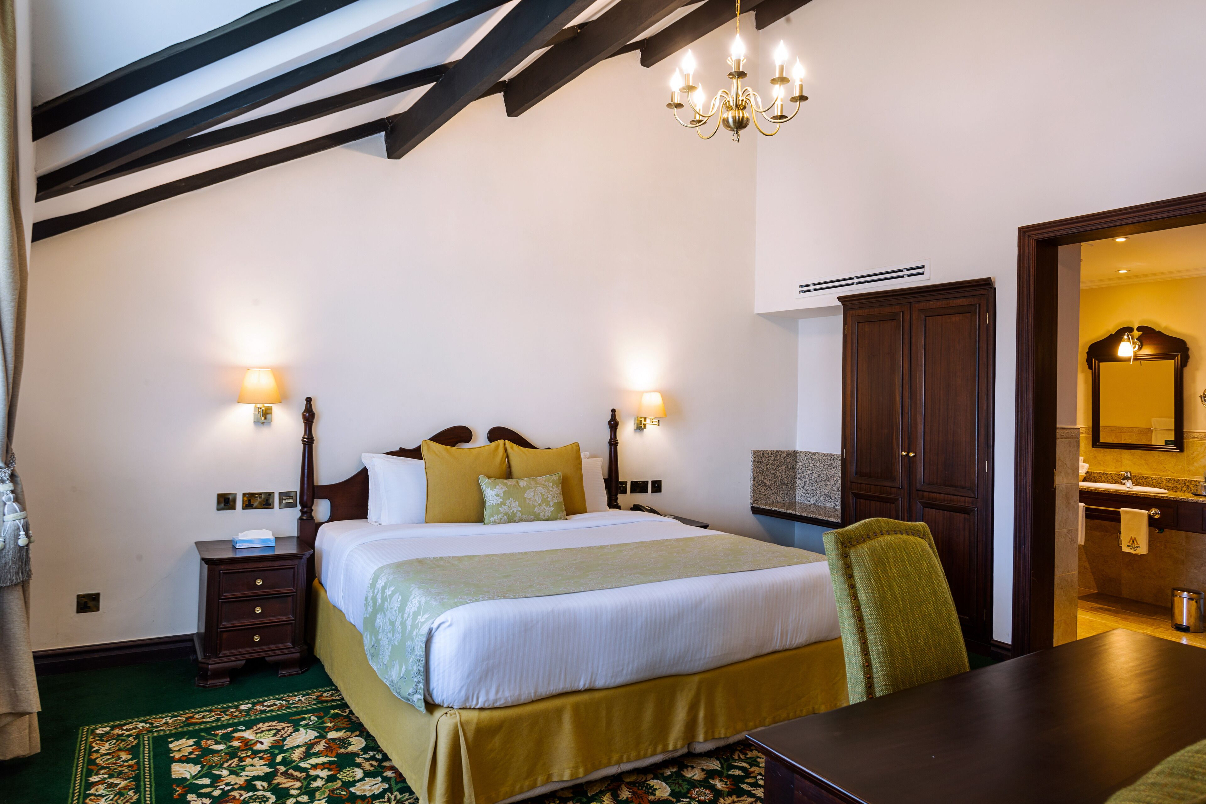 Habitación de hotel con una cama grande, dos lámparas de noche, una mesita de noche de madera, alfombra floral verde y una mesa de comedor de madera, además de un candelabro.