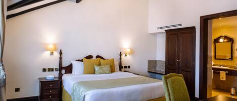 Habitación de hotel con una cama grande, dos lámparas de noche, una mesita de noche de madera, alfombra floral verde y una mesa de comedor de madera, además de un candelabro.