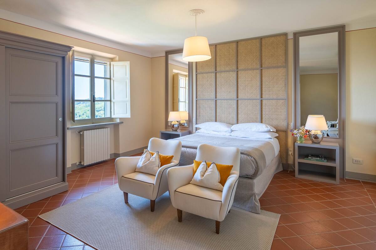 Quarto com cama grande, duas poltronas, mesa de cabeceira, espelho e janela com vista.