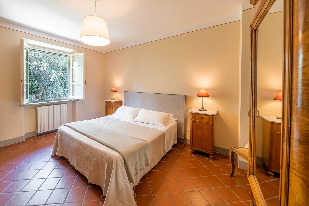 Quarto com cama, duas luminárias de cabeceira, janela e guarda-roupa de madeira.