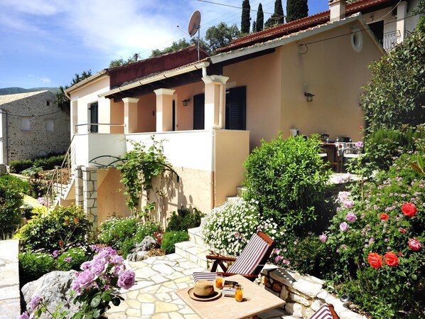 A house with a stone patio, a lounge chair, and a dining table with a hat and drinks.