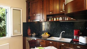 A kitchen with dark wooden cabinets, a black granite countertop, a sink, a red coffee maker, and a window with a view of greenery. - Villa Edem (Corfu)