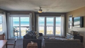 En stue med sofa, et lille bord og fjernsyn. Store vinduer med udsigt til stranden.