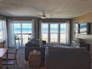 A living room with a sofa, a small table, and a television. There are large windows with a view of the beach. - Surf Side Six C-3 (St Augustine Beach)