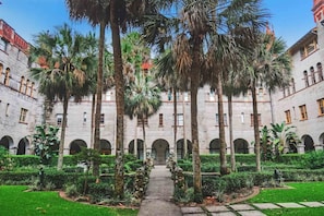 Un patio con un camino central bordeado por palmeras y un edificio con ventanas arqueadas.