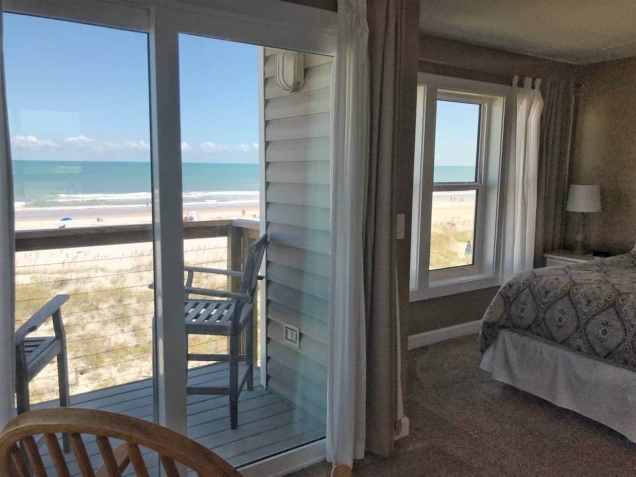 A room with a bed, a chair, and a view of the beach through a sliding door.