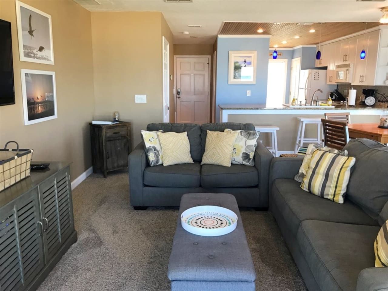 Ein Wohnzimmer mit grauem Sofa, grauem Hocker und Couchtisch. Im Hintergrund ist eine Küchenzeile erkennbar.