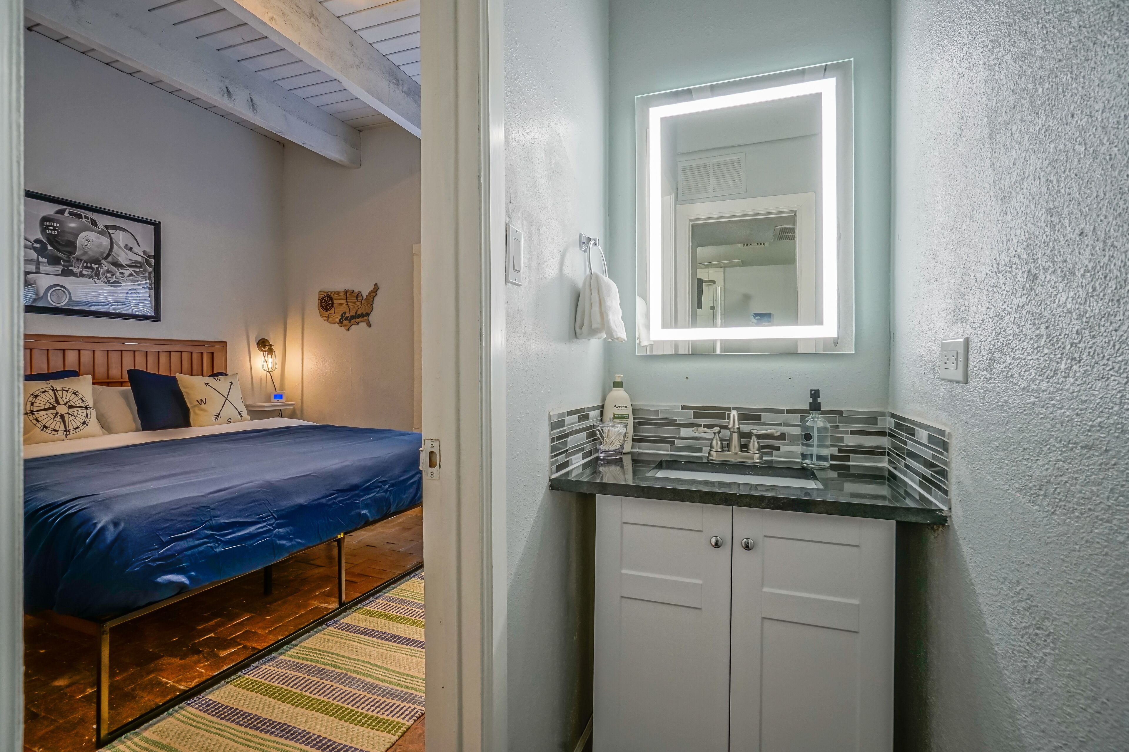 Un dormitorio con cama, mesita de noche, espejo con luz integrado en la pared y un baño pequeño con lavamanos y armarios.