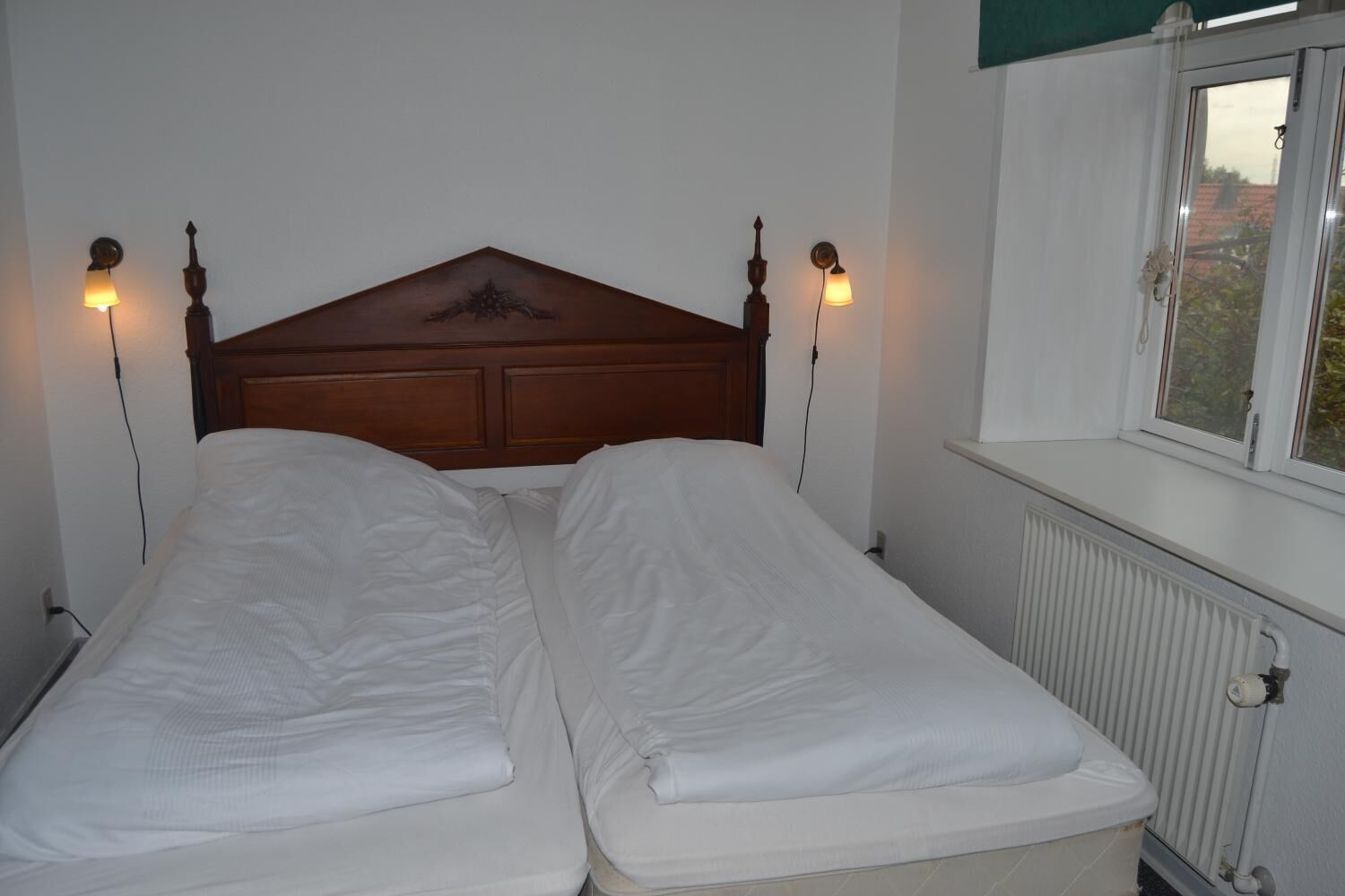 A double bed with a wooden headboard, two bedside lamps, and a window with a view of trees.