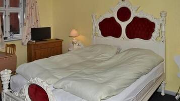 Una cama con cabecera blanca y detalles en rojo, un televisor sobre un aparador de madera y una lámpara en el aparador.