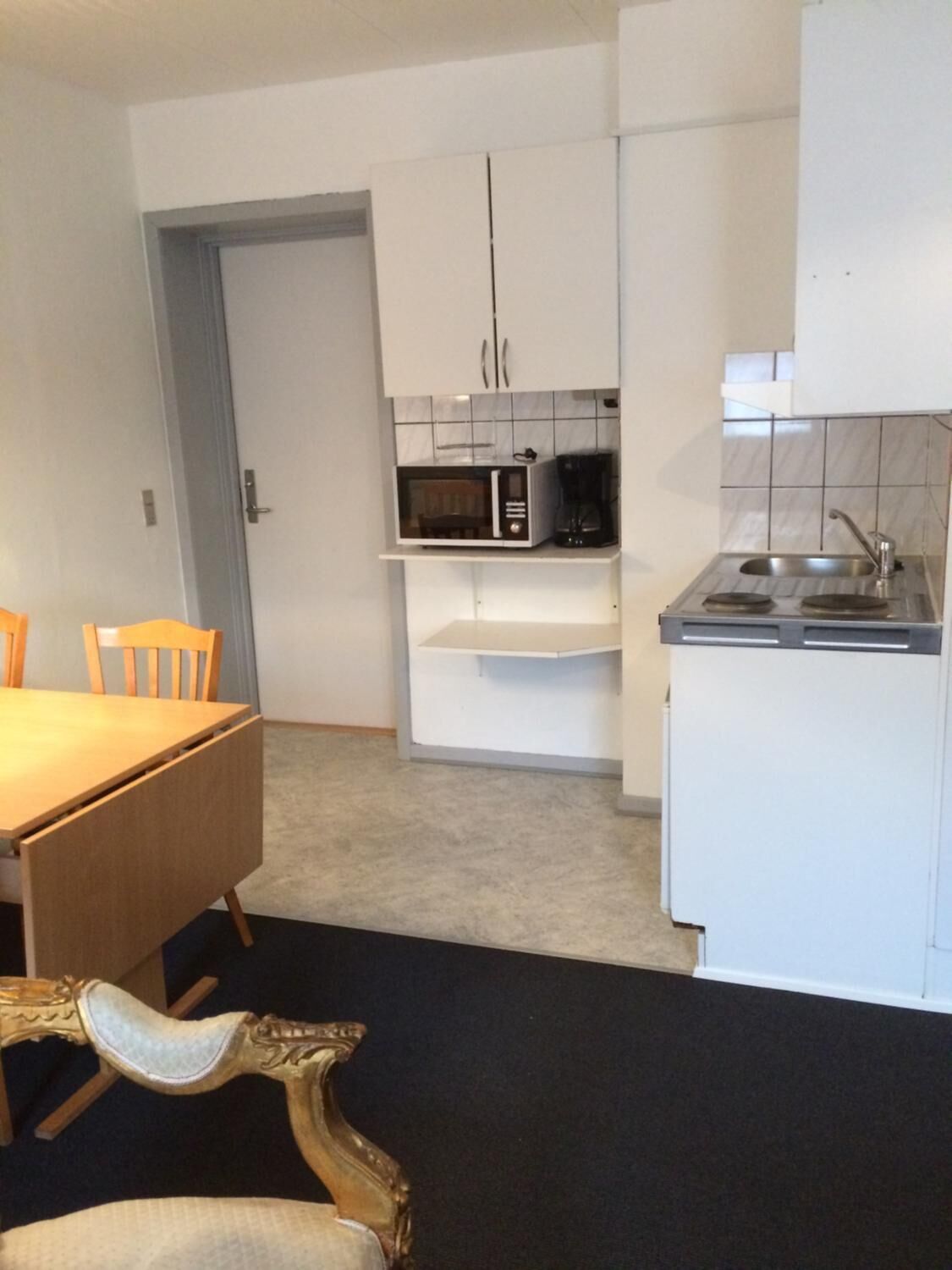 A compact kitchen with white cabinets, a microwave, a coffee maker, and a sink. There is a dining table with chairs and a decorative chair with a gold frame.