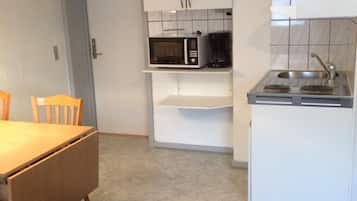 A compact kitchen with white cabinets, a microwave, a coffee maker, and a sink. There is a dining table with chairs and a decorative chair with a gold frame.