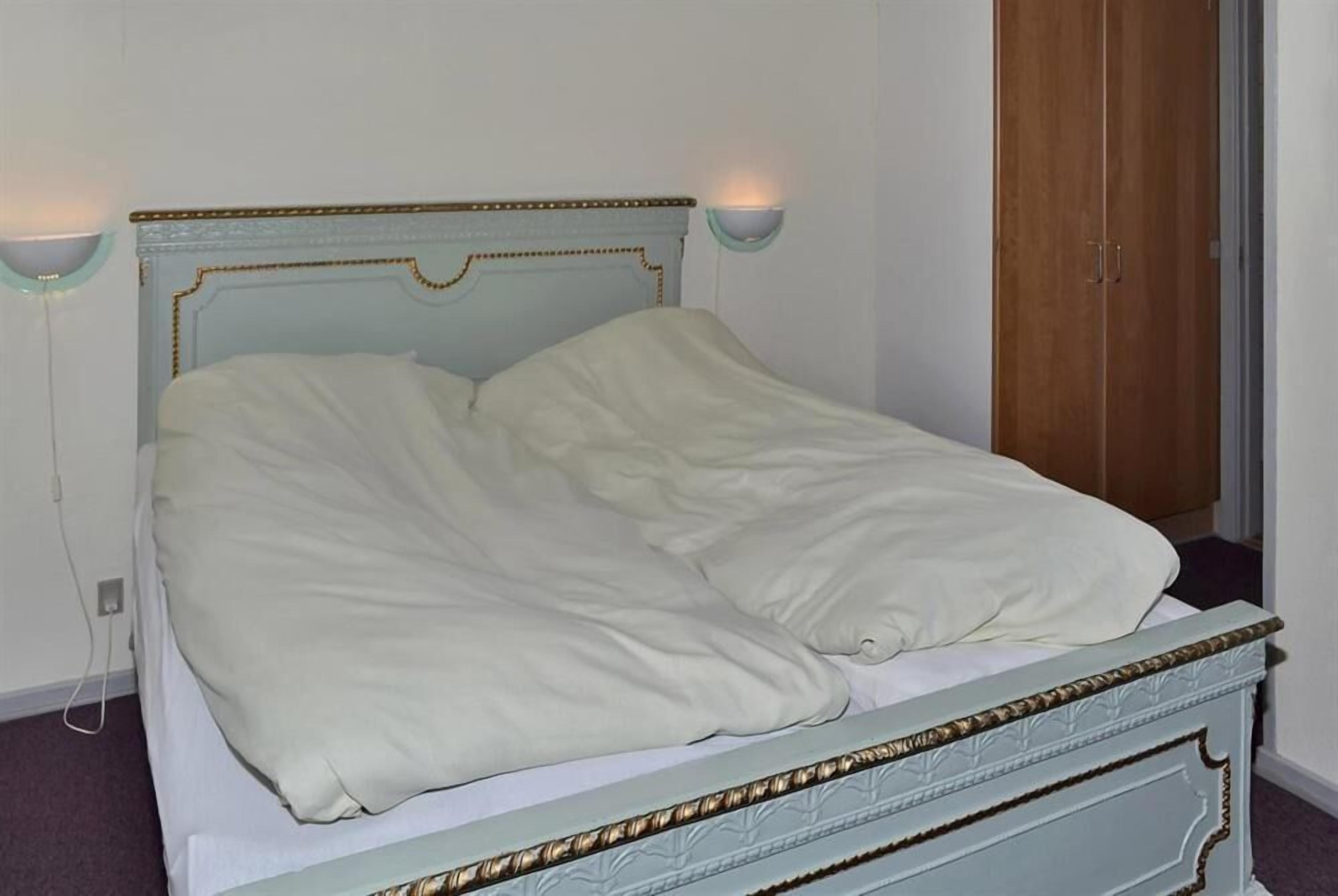 Una cama con cabecera azul claro y ropa de cama blanca en una habitación con un armario de madera y una lámpara fijada a la pared.