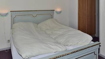 Una cama con cabecera azul claro y ropa de cama blanca en una habitación con un armario de madera y una lámpara fijada a la pared.