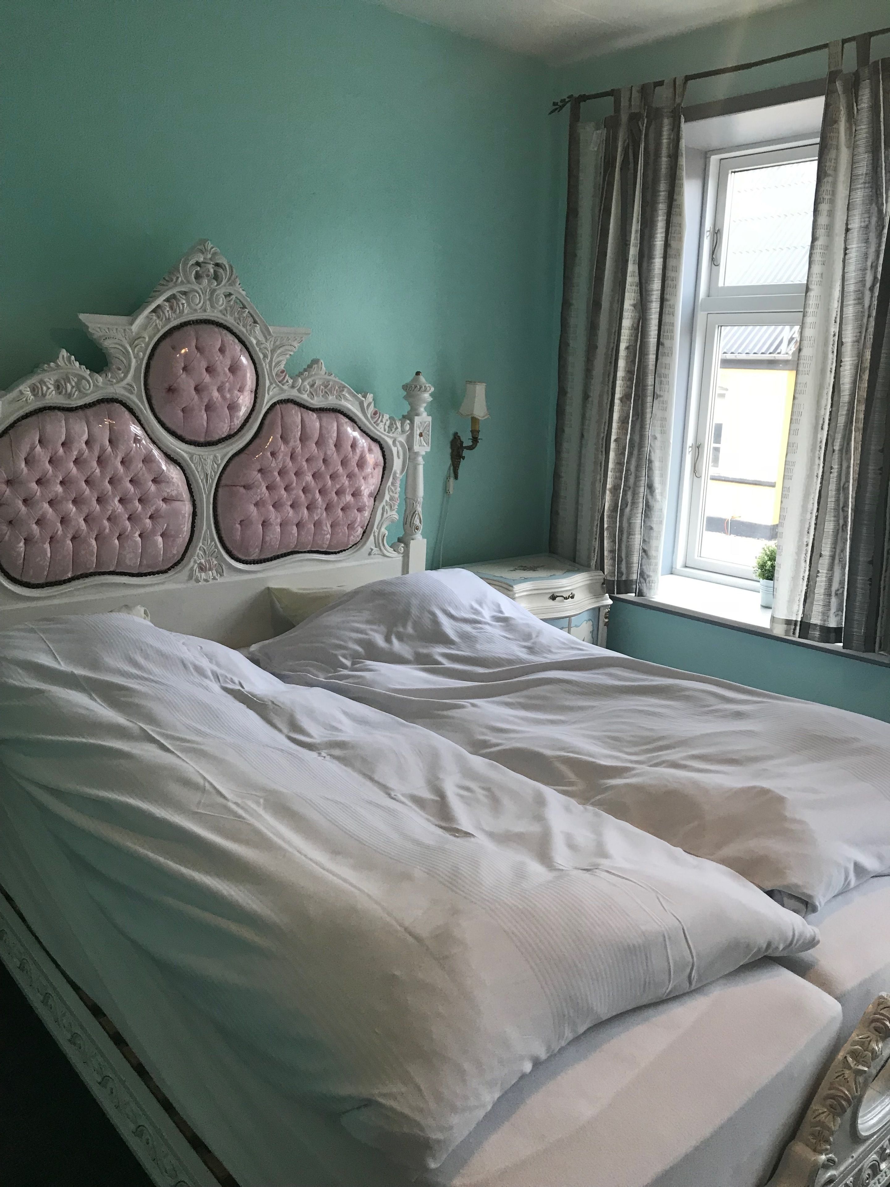 Un letto con testiera imbottita, un comodino con una lampada e una finestra con tende.