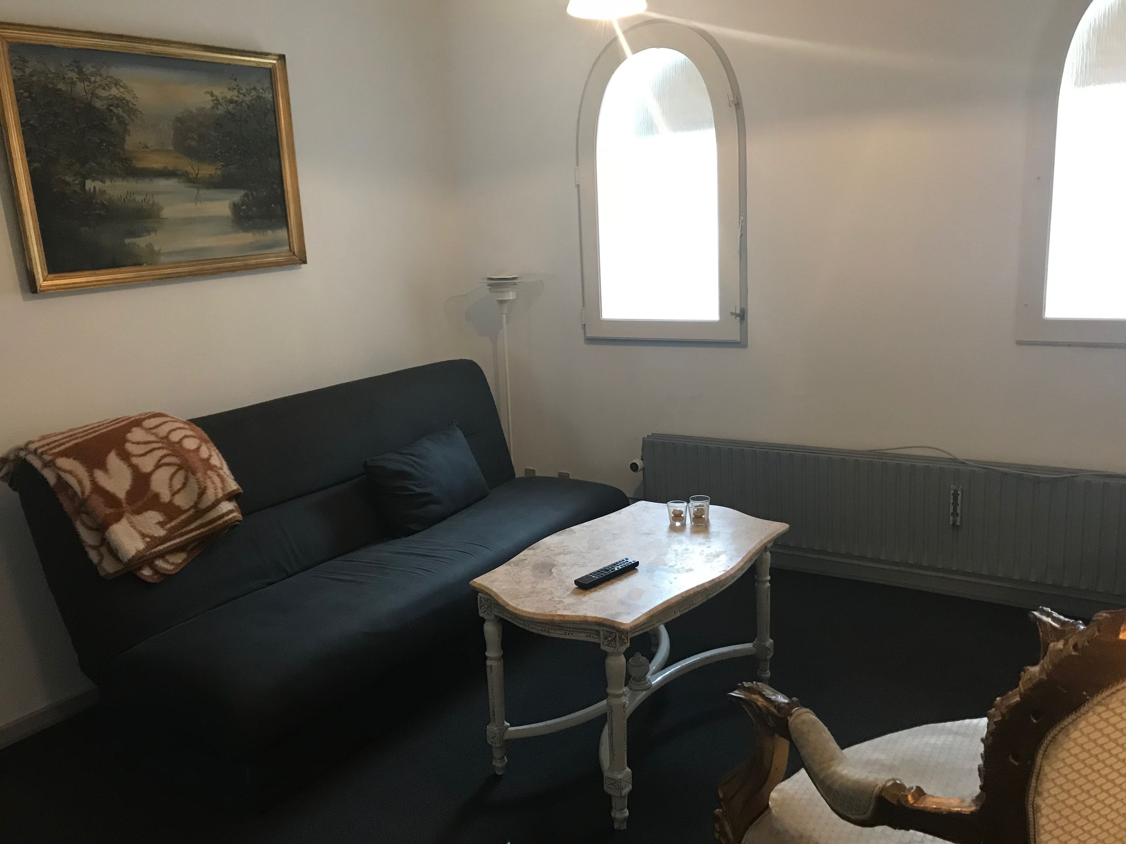 Una habitación con un sofá azul oscuro, una pequeña mesa blanca con un control remoto y una silla decorativa con respaldo tallado.