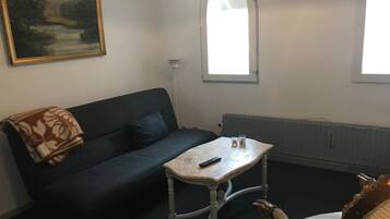 Una habitación con un sofá azul oscuro, una pequeña mesa blanca con un control remoto y una silla decorativa con respaldo tallado.