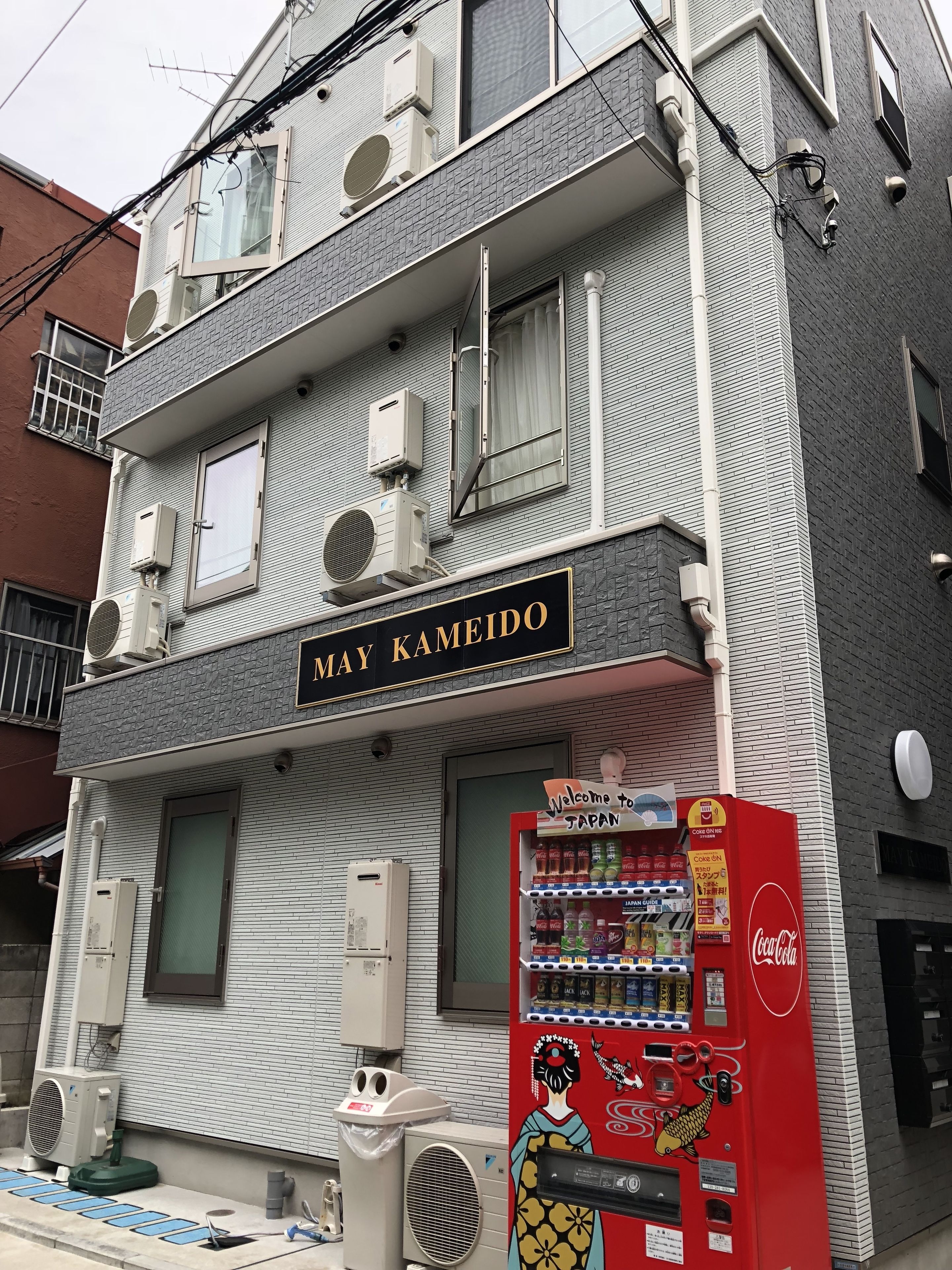 Een gebouw met een bord waarop 'May Kameido' staat en een automaat met Japanse en Coca-Cola producten.