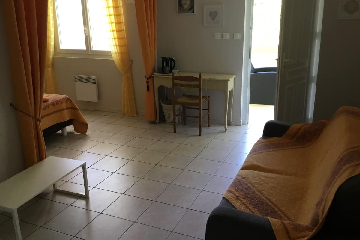 Une pièce comprenant un lit, un bureau, une chaise et un banc. On y trouve une fenêtre avec des rideaux, un radiateur et une porte.