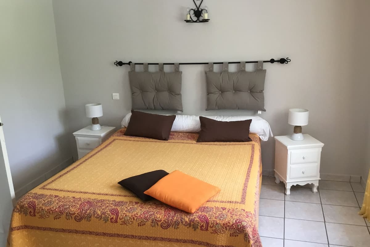 Une chambre bien rangée, avec un lit, deux tables de chevet, une petite commode, un luminaire fixé au mur et une petite étagère.