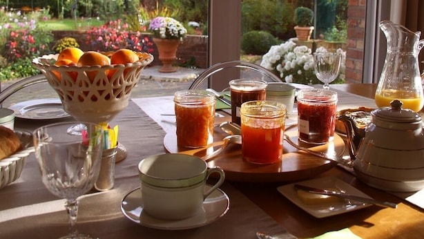 En frukostuppsättning med ett varierat utbud av mat och dryck, inklusive en fruktskål, burkar med sylt, en tekanna och en kanna med apelsinjuice.