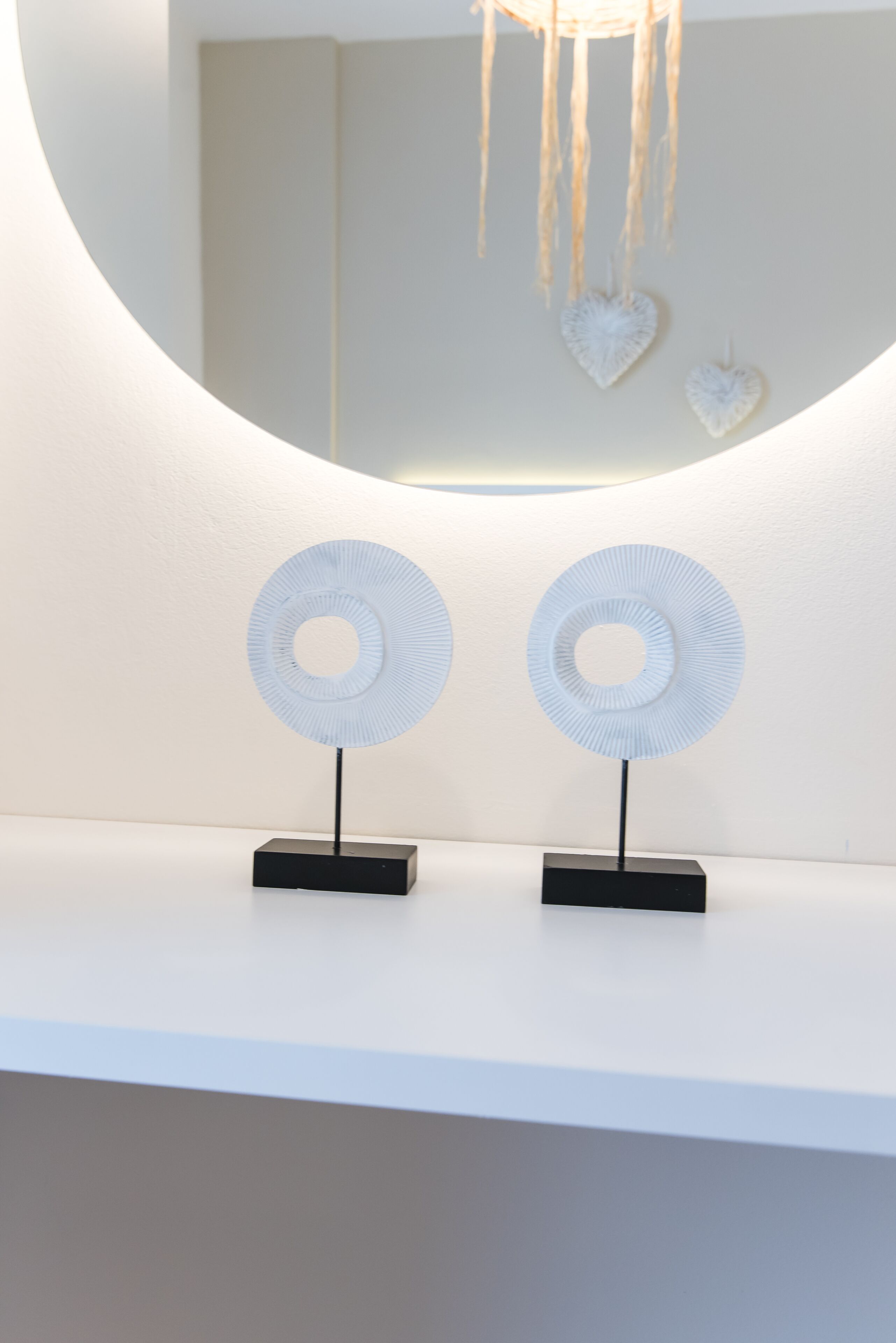 Deux ventilateurs blancs, de forme circulaire et indépendants, sont posés sur une surface blanche. Un miroir rond reflète un luminaire suspendu et deux décorations en forme de cœur.
