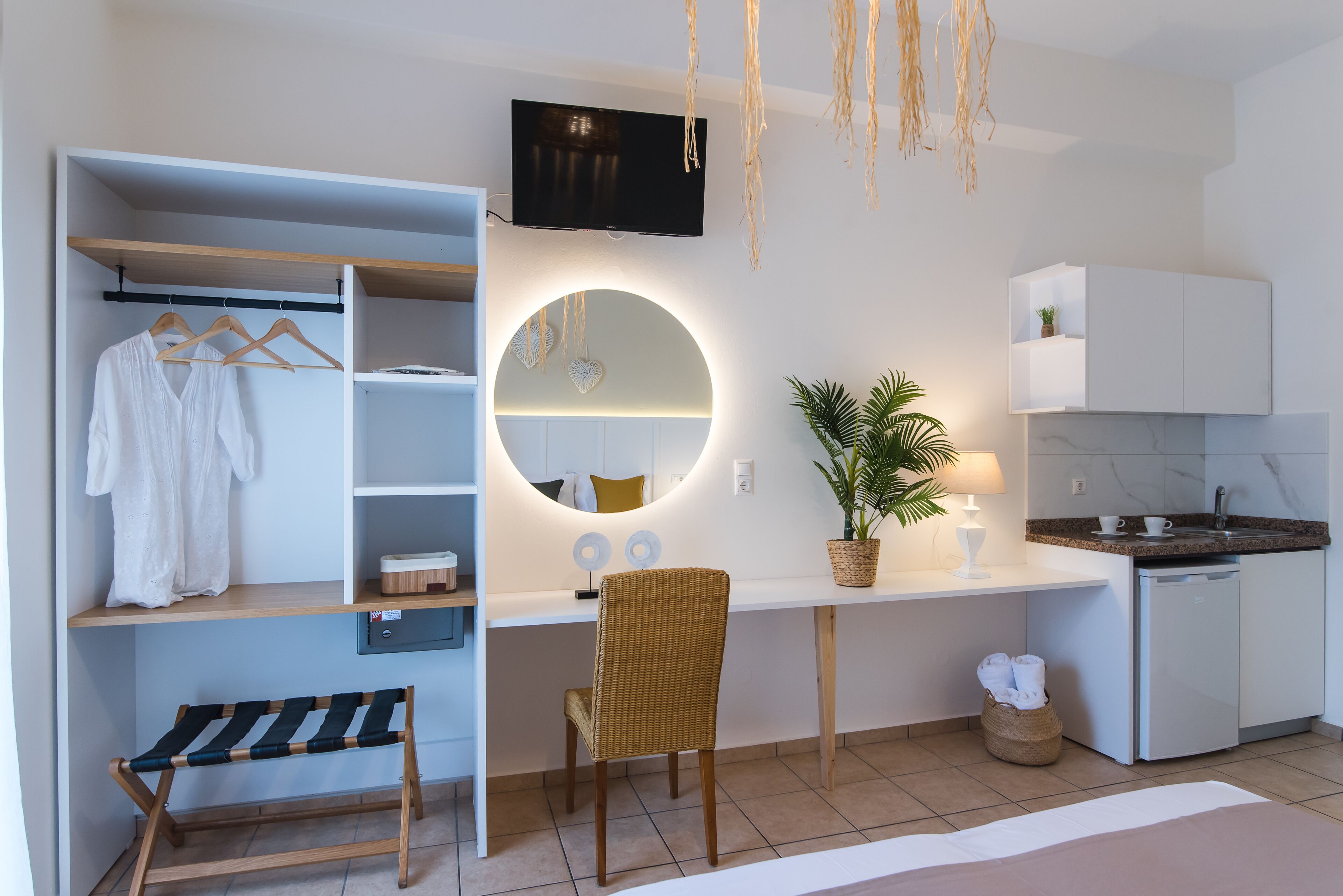 A modern, well-lit room with a wardrobe, a desk with a TV, a chair, a plant, and a small kitchen area.