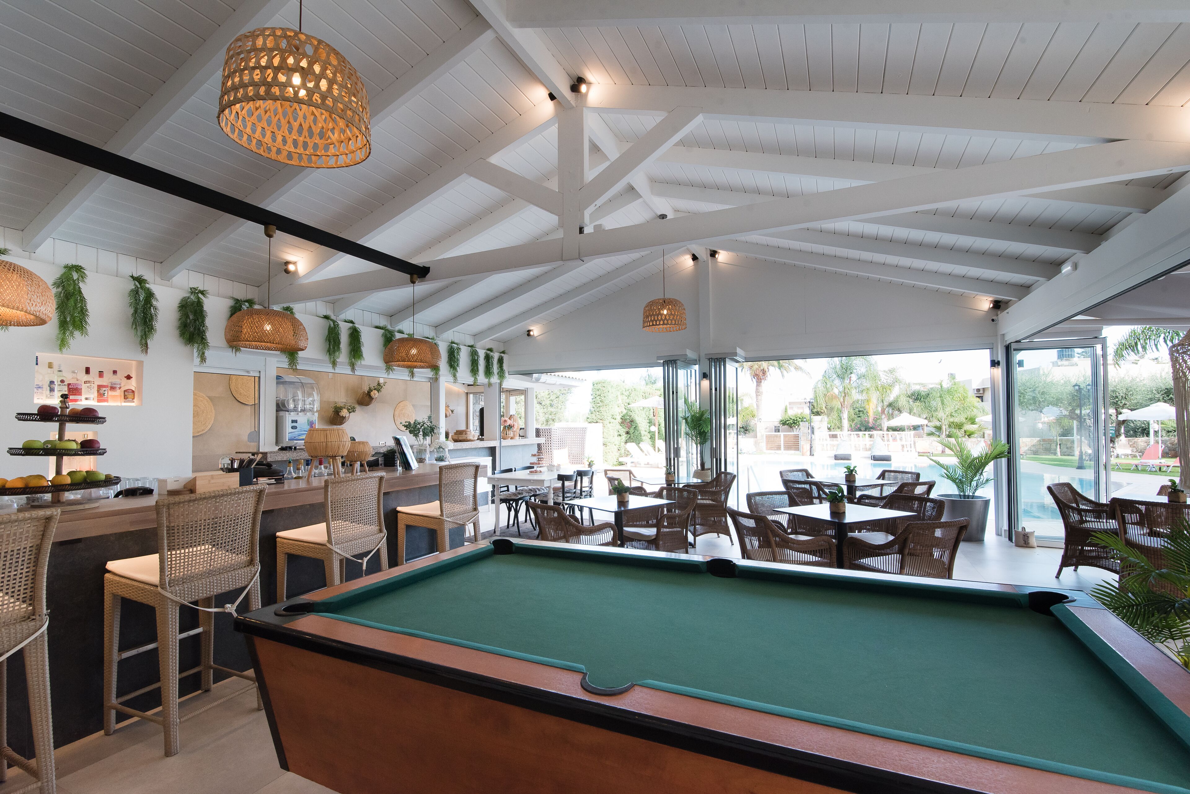 A pool table in a room with wicker chairs and hanging lamps. There are tables and chairs outside the window.