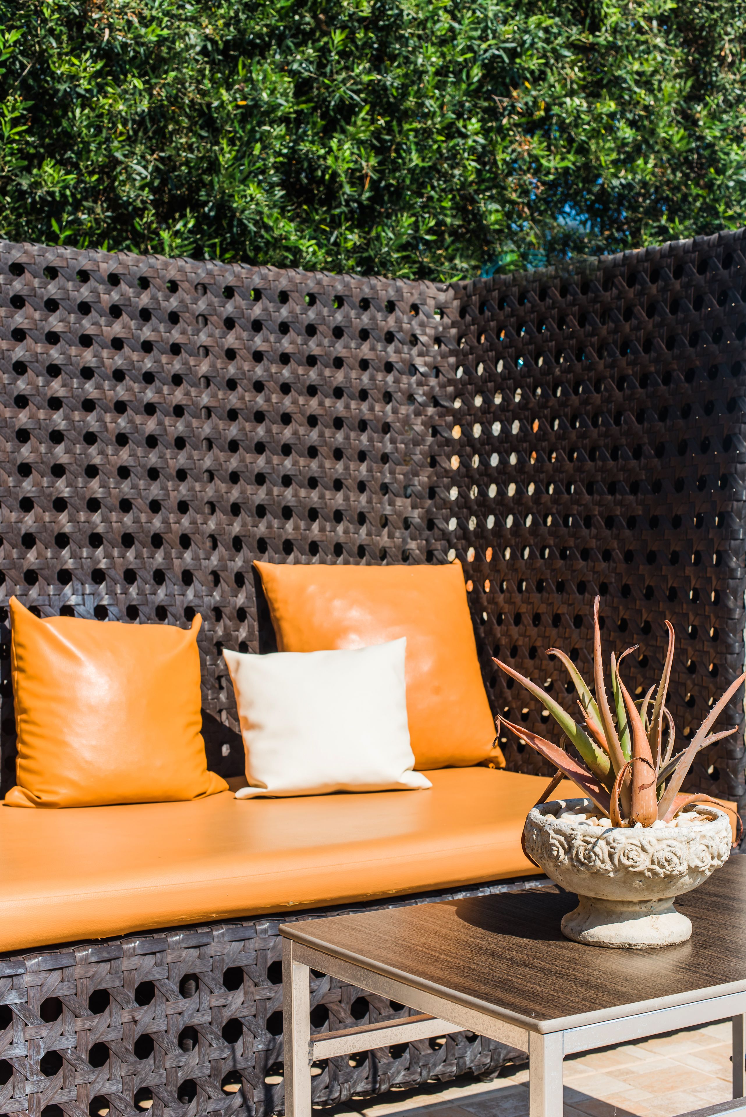 A modern outdoor seating area with a woven backrest, two orange cushions, a white cushion, and a potted plant on a table.