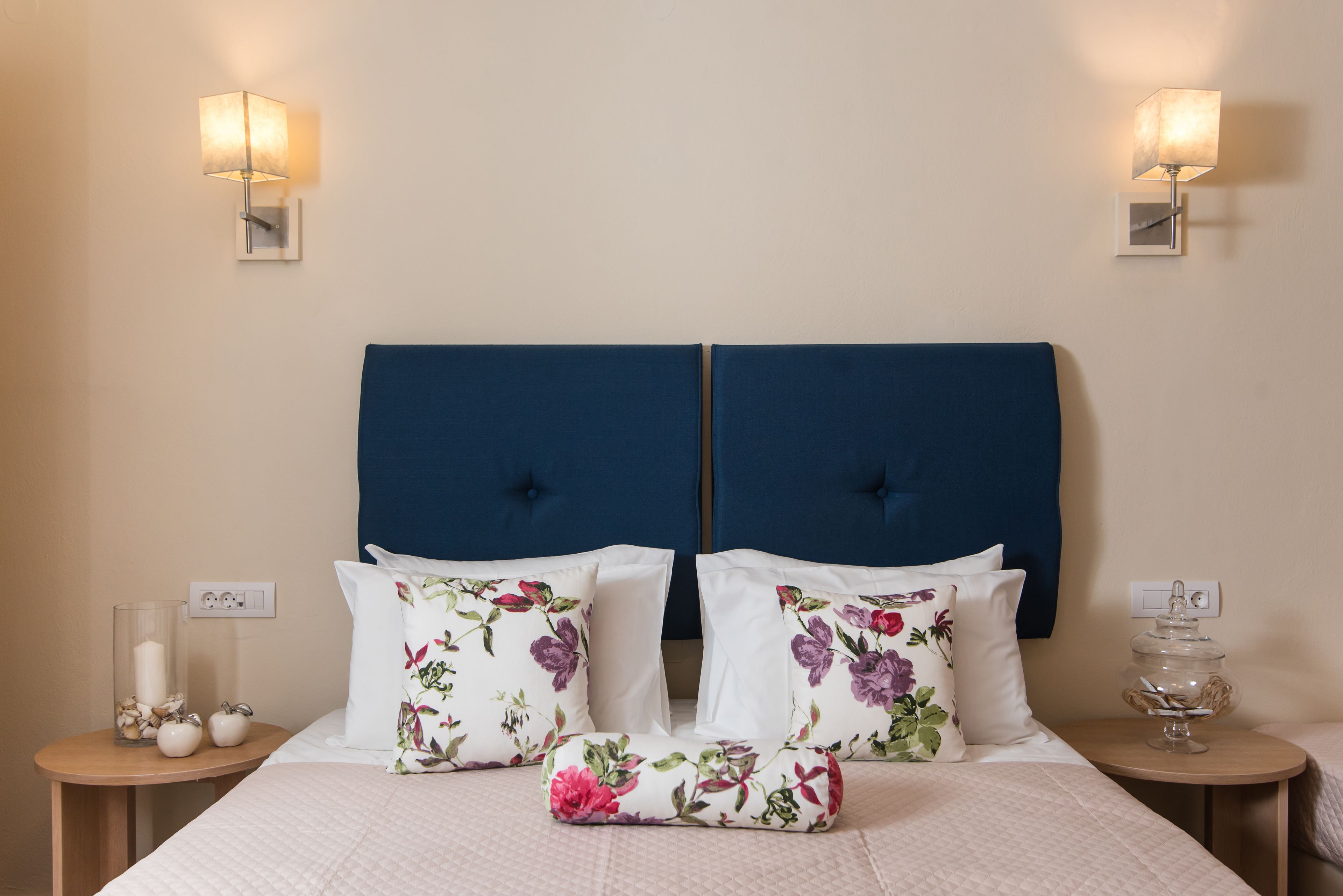 A hotel room with a large bed, floral pillows, two bedside tables with lamps, and a nightstand with a candle and decorative items.