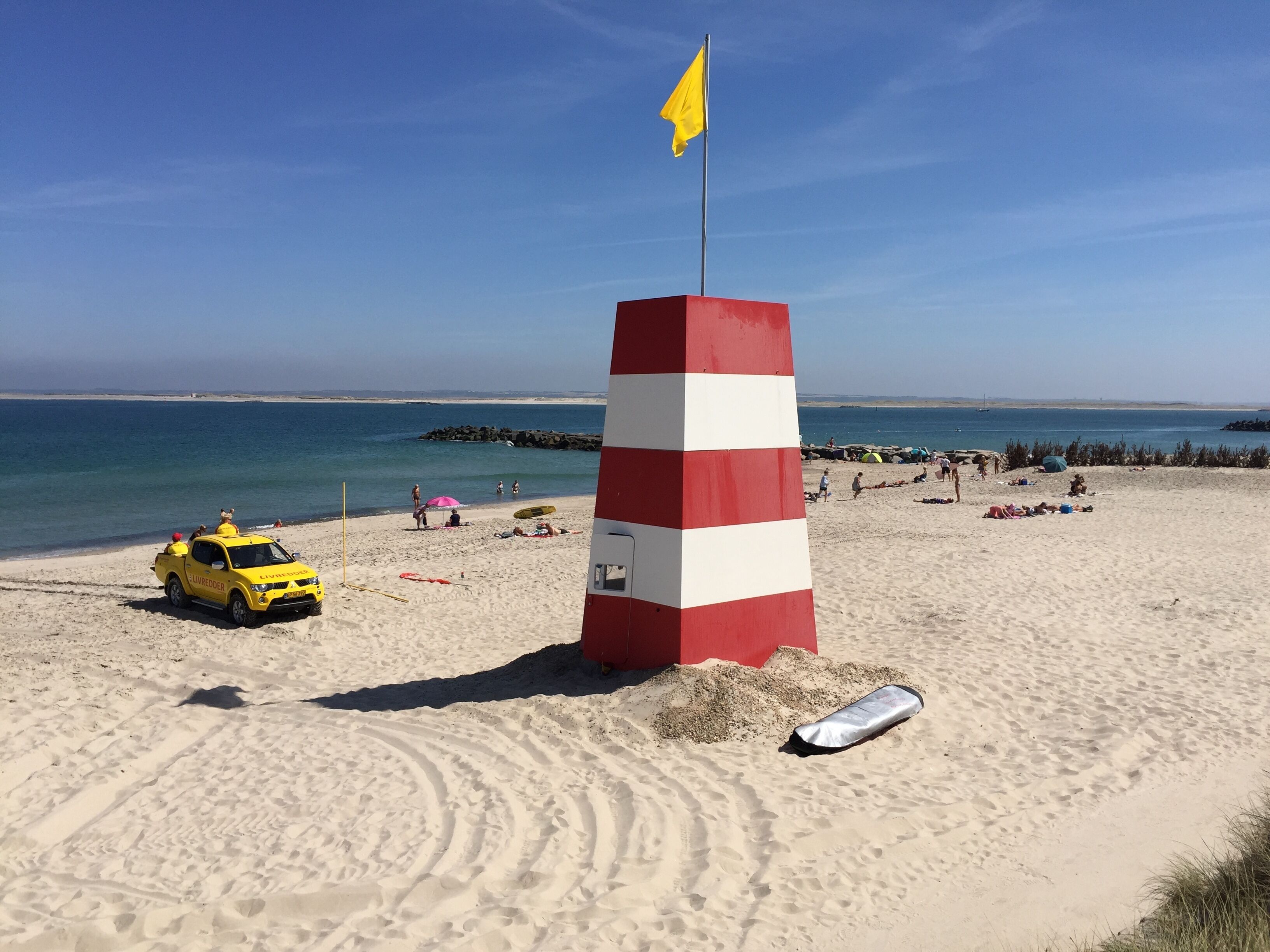 A beach with a red and white striped lighthouse, a yellow lifeguard truck, and people enjoying the beach.
