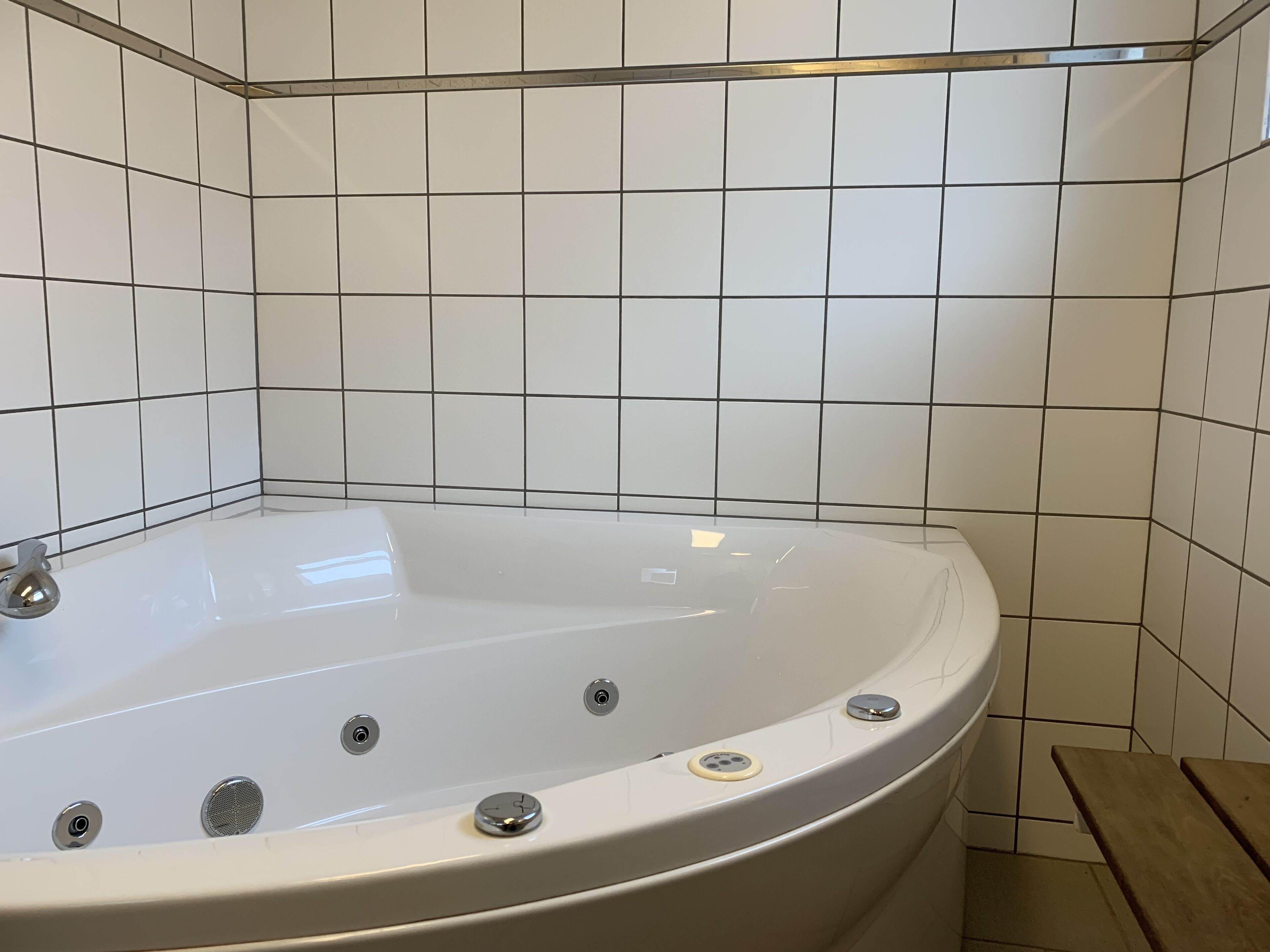 A corner bathtub with a wooden bench, white tiled walls, and a metal shelf above the tub.