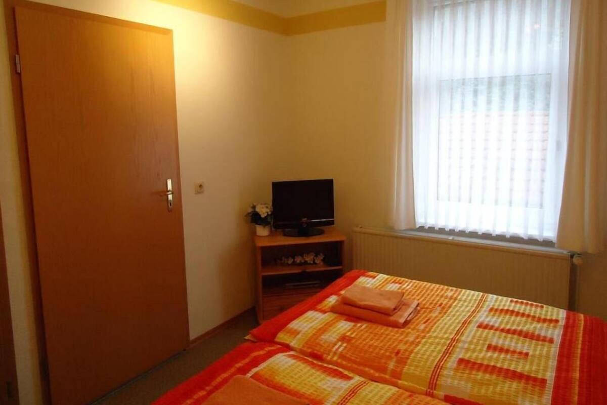 Une chambre à coucher avec un lit, un téléviseur posé sur un meuble, une fenêtre avec des rideaux et une porte.