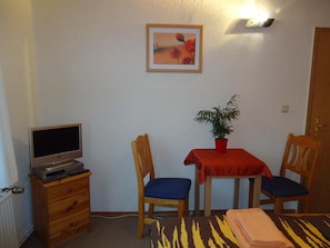 A small room with a desk, two chairs, a table with a red tablecloth, a potted plant, a television on a wooden cabinet, and a framed picture on the wall. - Pension Thaler Hof (Thale)
