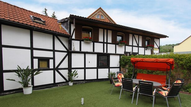 A traditional half-timbered house with a red roof, a patio with artificial grass, and outdoor furniture. - Pension Thaler Hof (Thale)
