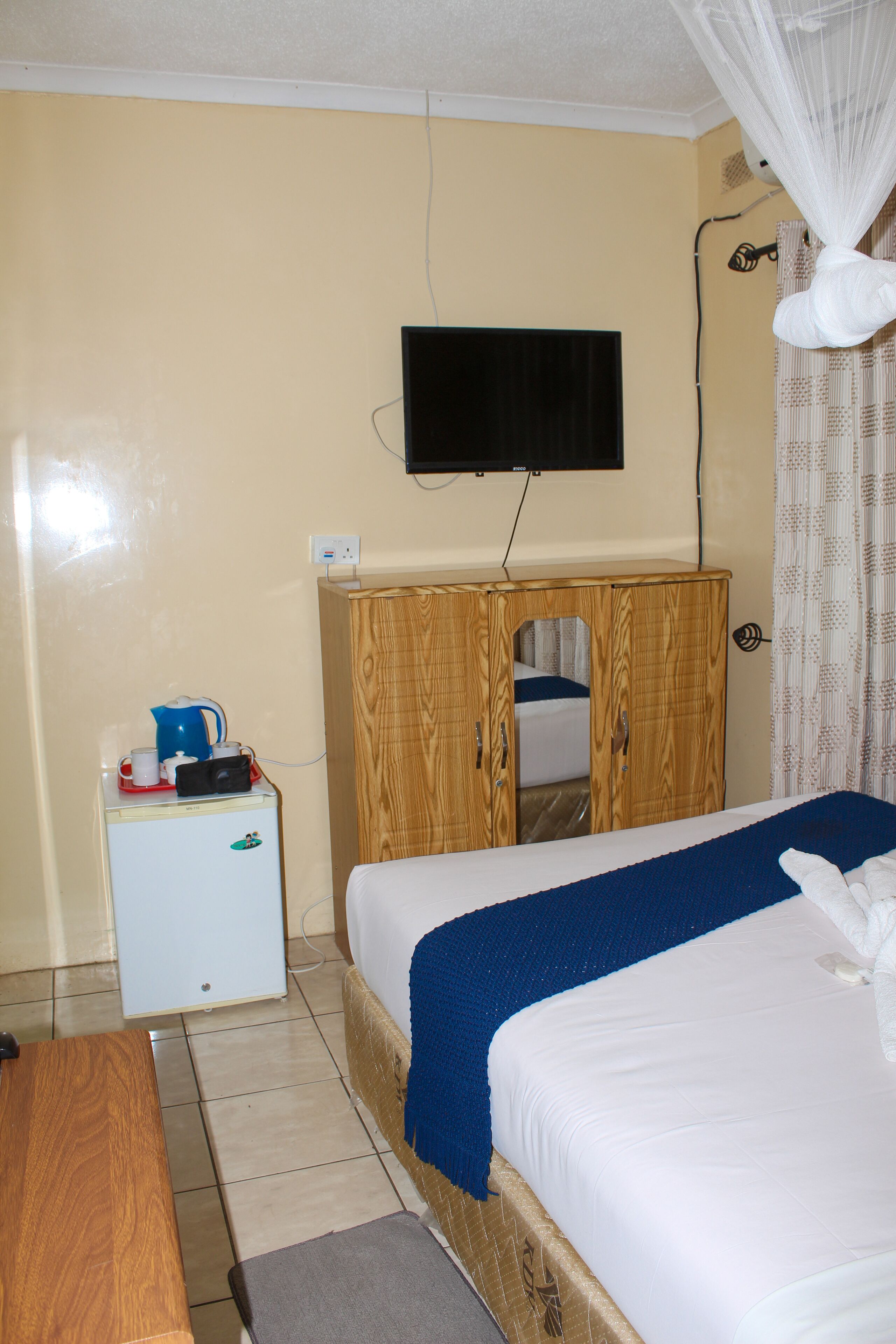 Un dormitorio con cama, un armario de madera, un refrigerador blanco pequeño, un televisor montado en la pared y una mosquitera sobre la cama.