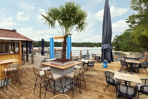 An outdoor dining area with wooden tables and chairs, a central planter with a palm tree, and umbrellas providing shade.