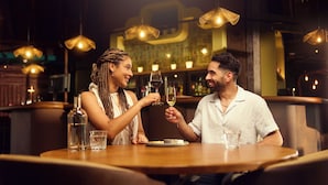 Un couple est assis à une table en bois dans un restaurant, avec des lampes suspendues et un bar en arrière-plan.