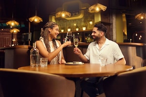 A couple sitting at a wooden table in a restaurant with hanging lights and a bar in the background.