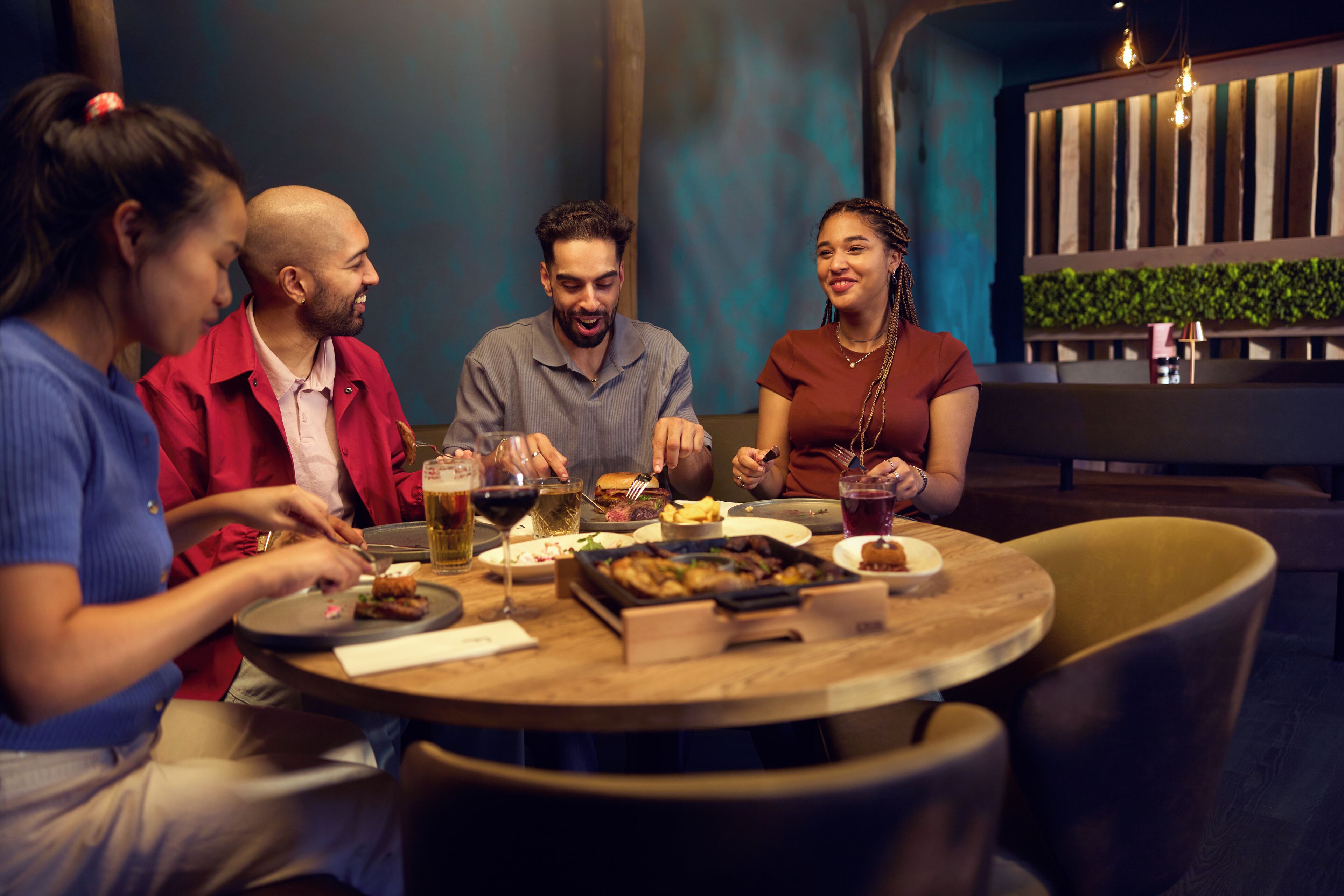 Een groep mensen dineert aan een ronde tafel in een restaurant met een modern interieur, inclusief een houten tafel en stoelen, en een decoratieve wand met verticale houten lamellen en groen.