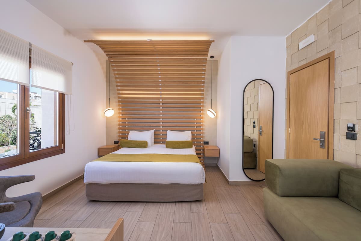 Una habitación de hotel moderna con una cama grande, mesitas de noche, un espejo, un sofá y vistas al exterior.