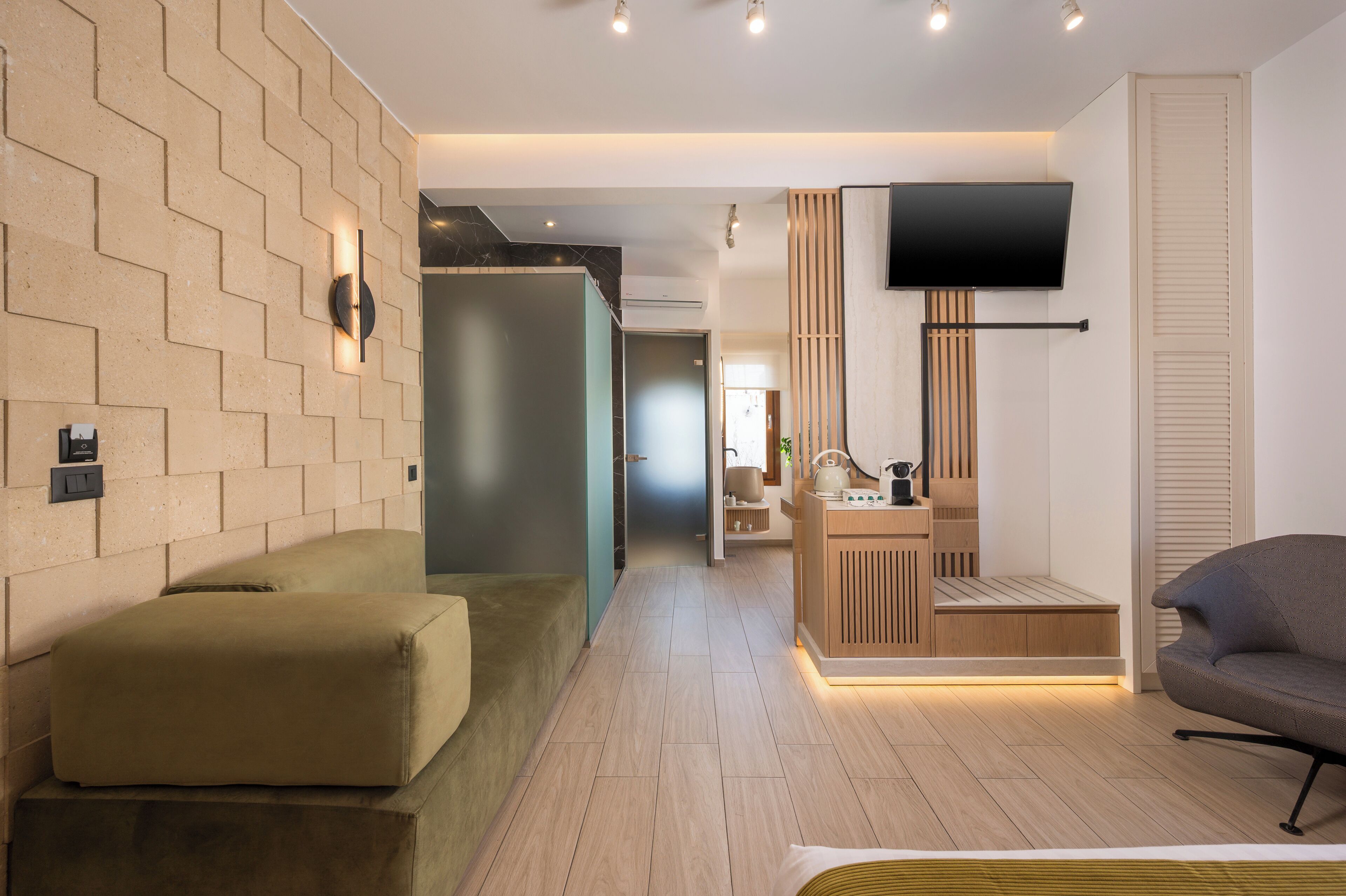 Quarto de hotel moderno com piso de madeira, um sofá, uma TV fixada na parede e um armário de madeira com um banquinho.