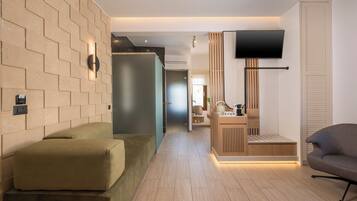 A modern hotel room with a wooden floor, a sofa, a TV mounted on the wall, and a wooden cabinet with a bench.