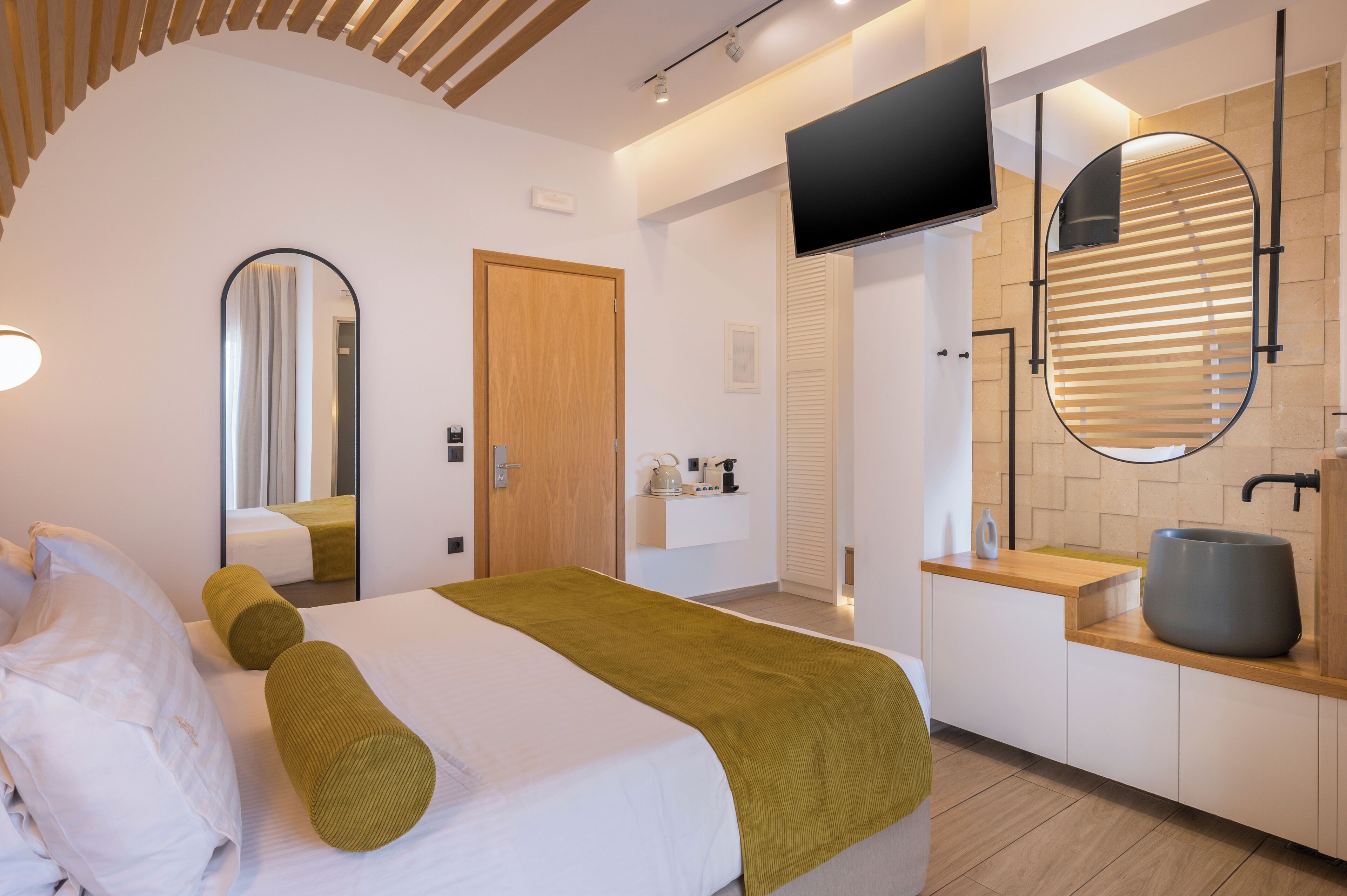 Una habitación de hotel moderna con cama, televisor, baño con lavamanos y espejo, y techo de madera.