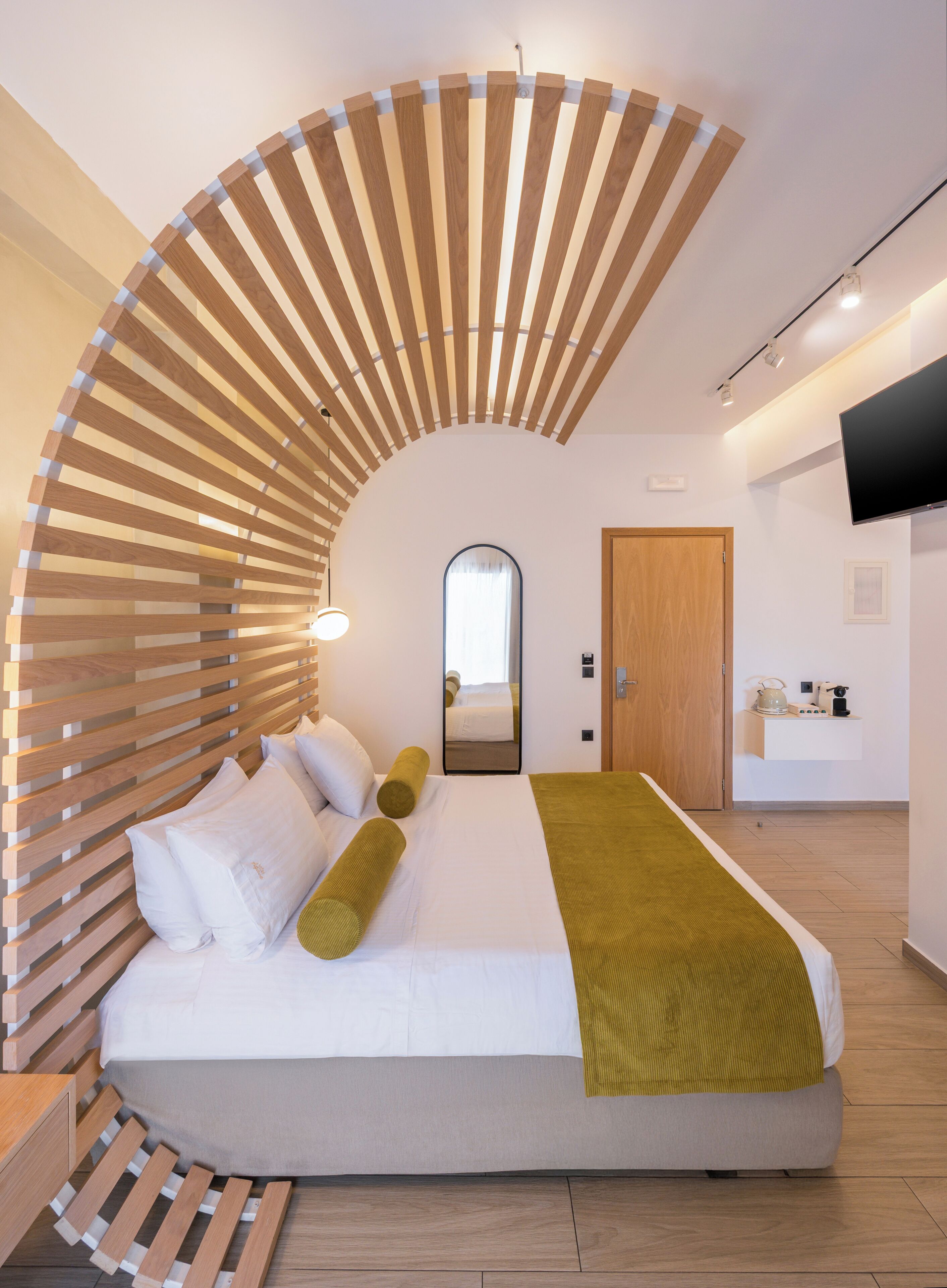 A modern hotel room with a curved wooden headboard, a large bed with white bedding and a green throw, a flat-screen TV mounted on the wall, and a small kitchenette area.