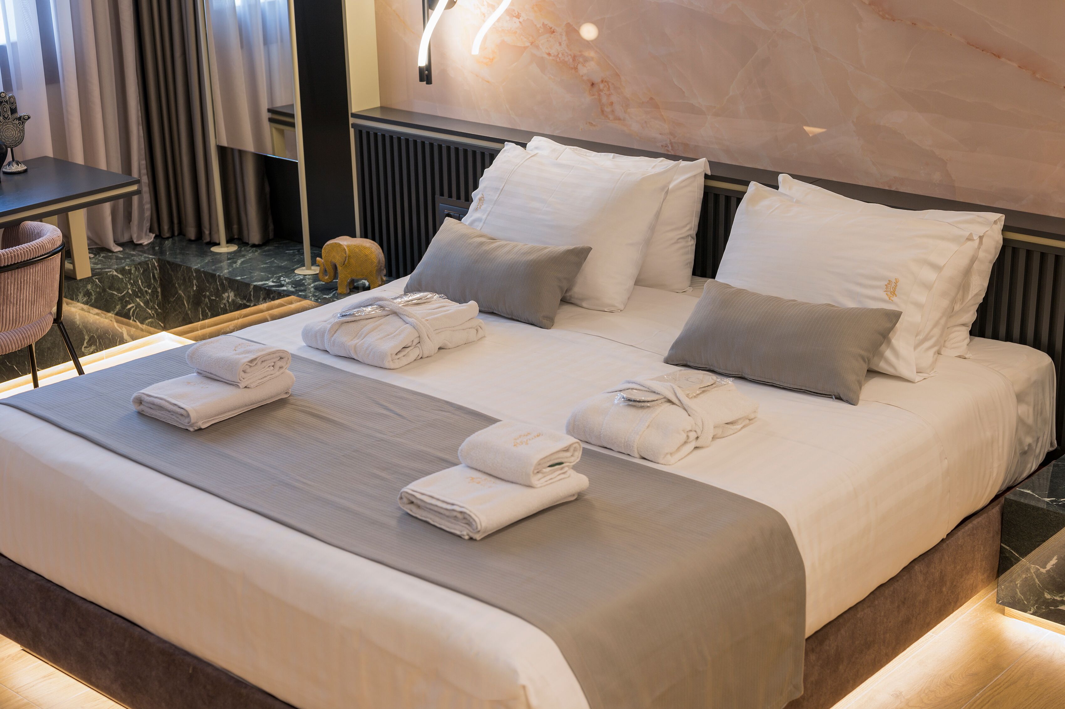 A neatly made bed with white linens and pillows, a small table with a chair, and a marble wall with a light fixture.