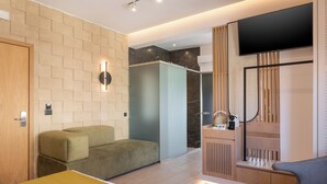 Ein modernes Hotelzimmer mit einer Couch, einem kleinen Tisch, einem an der Wand montierten Fernseher und einem Holzschrank mit Spiegel.