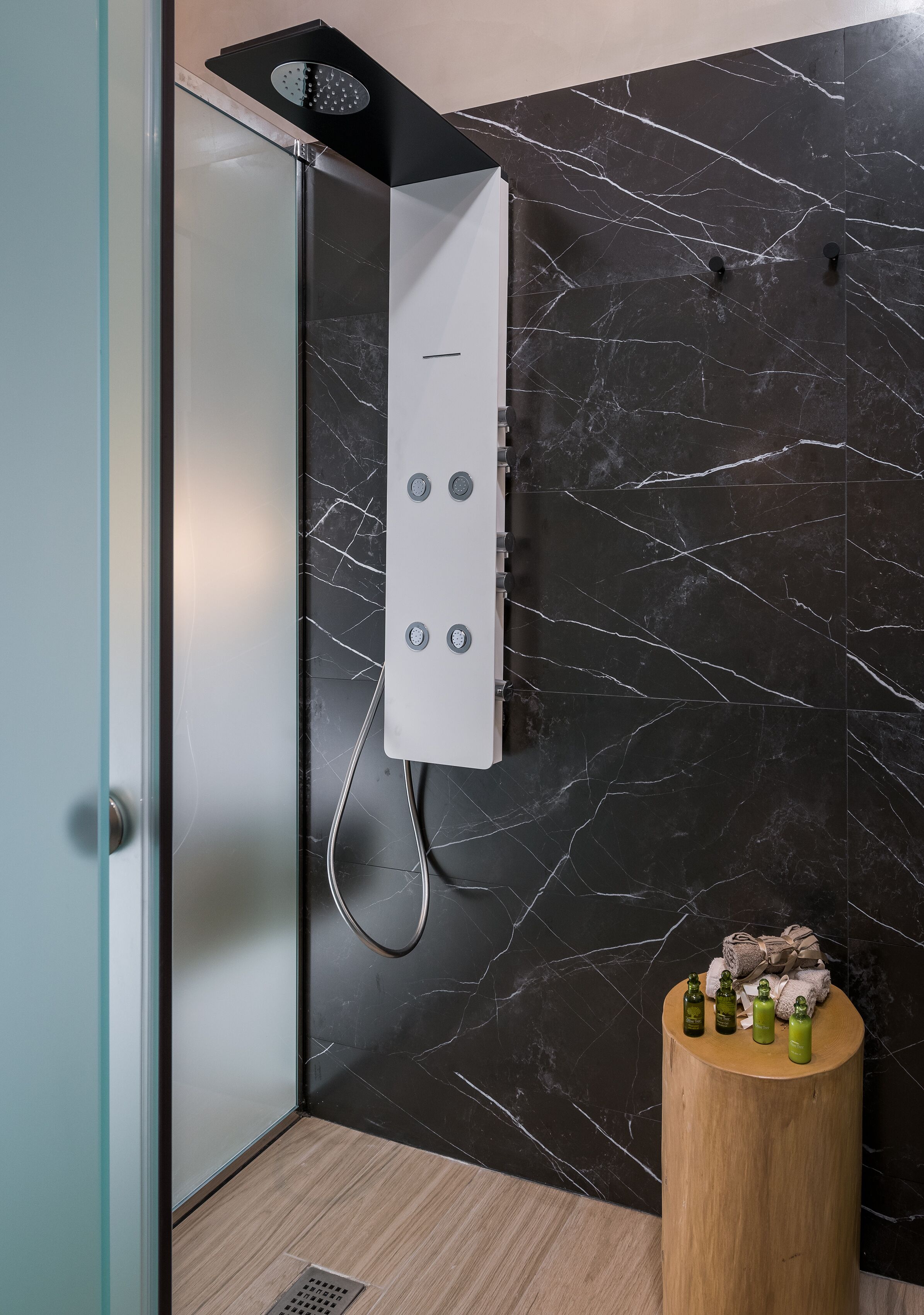 Un baño moderno con ducha, paredes de mármol y un taburete de madera con botellas.