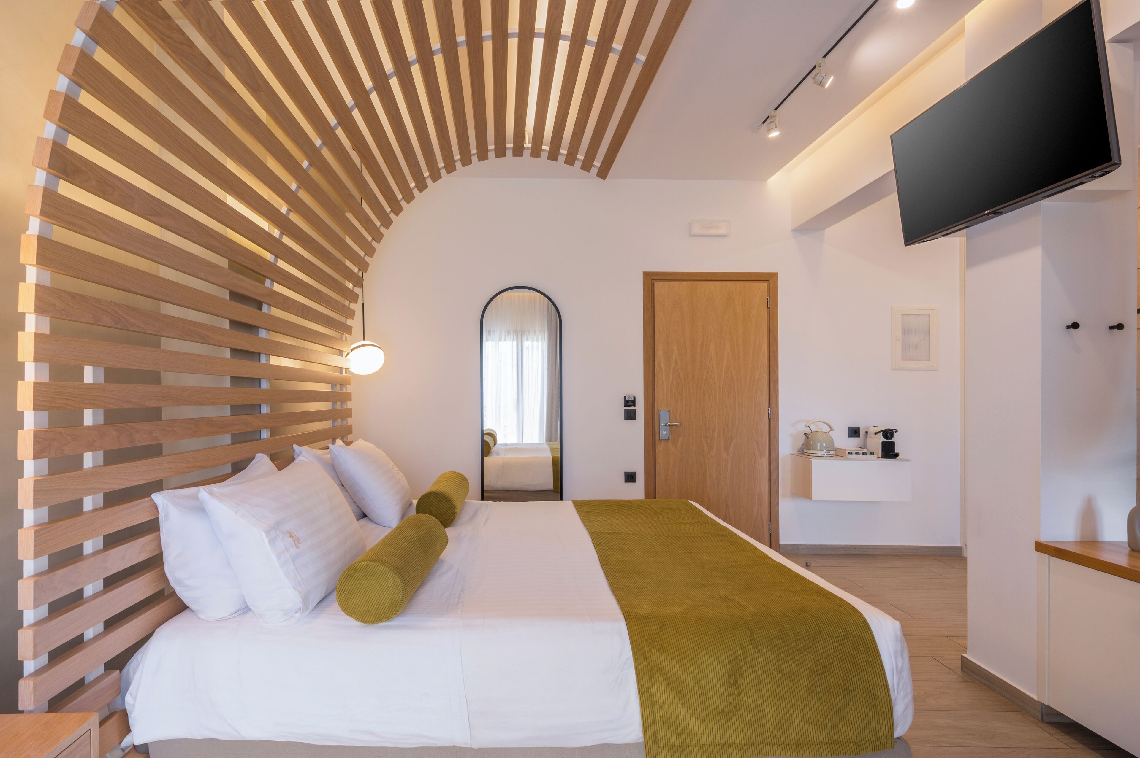 Une chambre d’hôtel moderne avec une tête de lit en lattes de bois, un grand lit avec une literie blanche et verte, un téléviseur à écran plat fixé au mur et un petit coin cuisine.