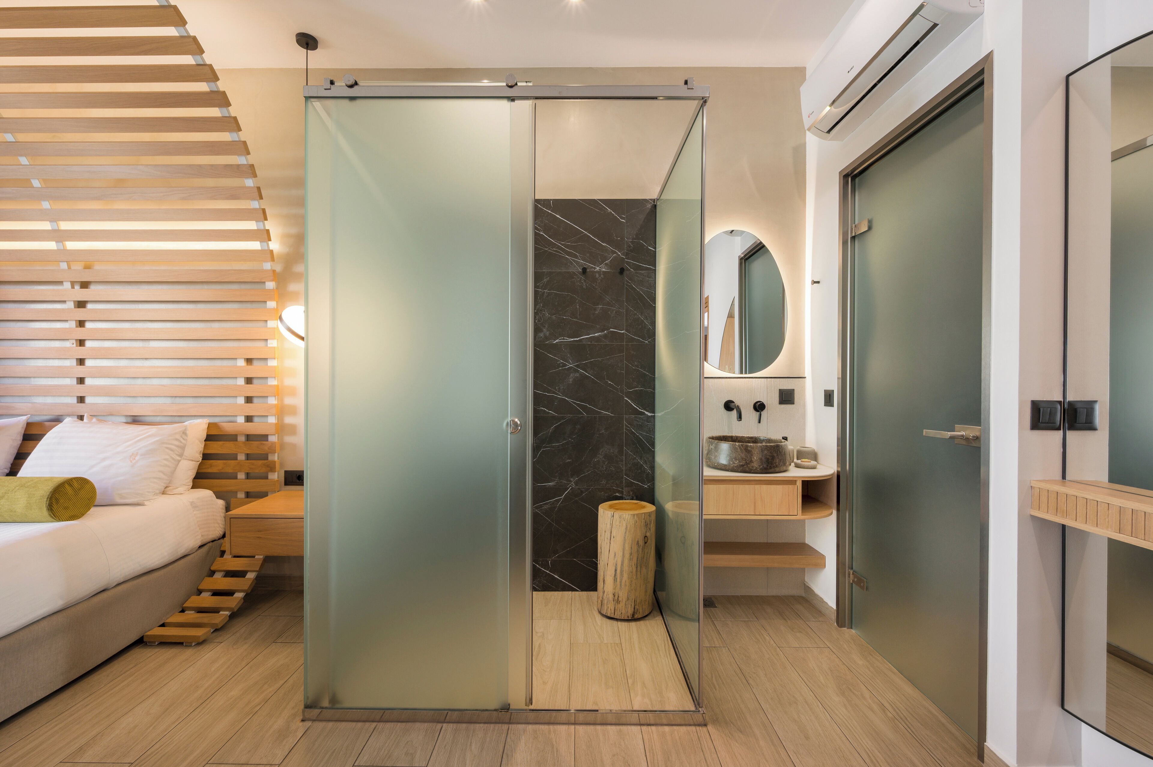 A modern bedroom with a bed, a wooden headboard, a sliding glass door leading to a bathroom, and a mirror.